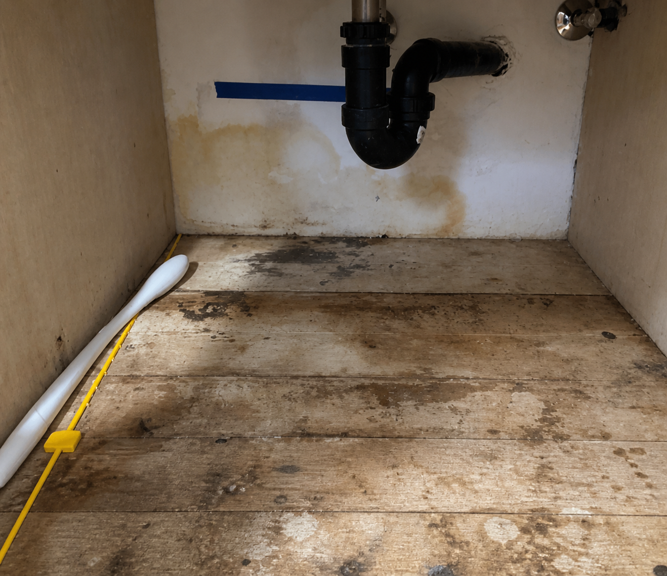 Under-sink plumbing with black pipes, a white pipe, and a yellow drain snake on a wooden floor, in a space with stained white walls.