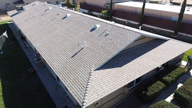 Aerial view of a house with a light gray roof, multiple vents, a porch, and landscaped yard with bushes and trees.