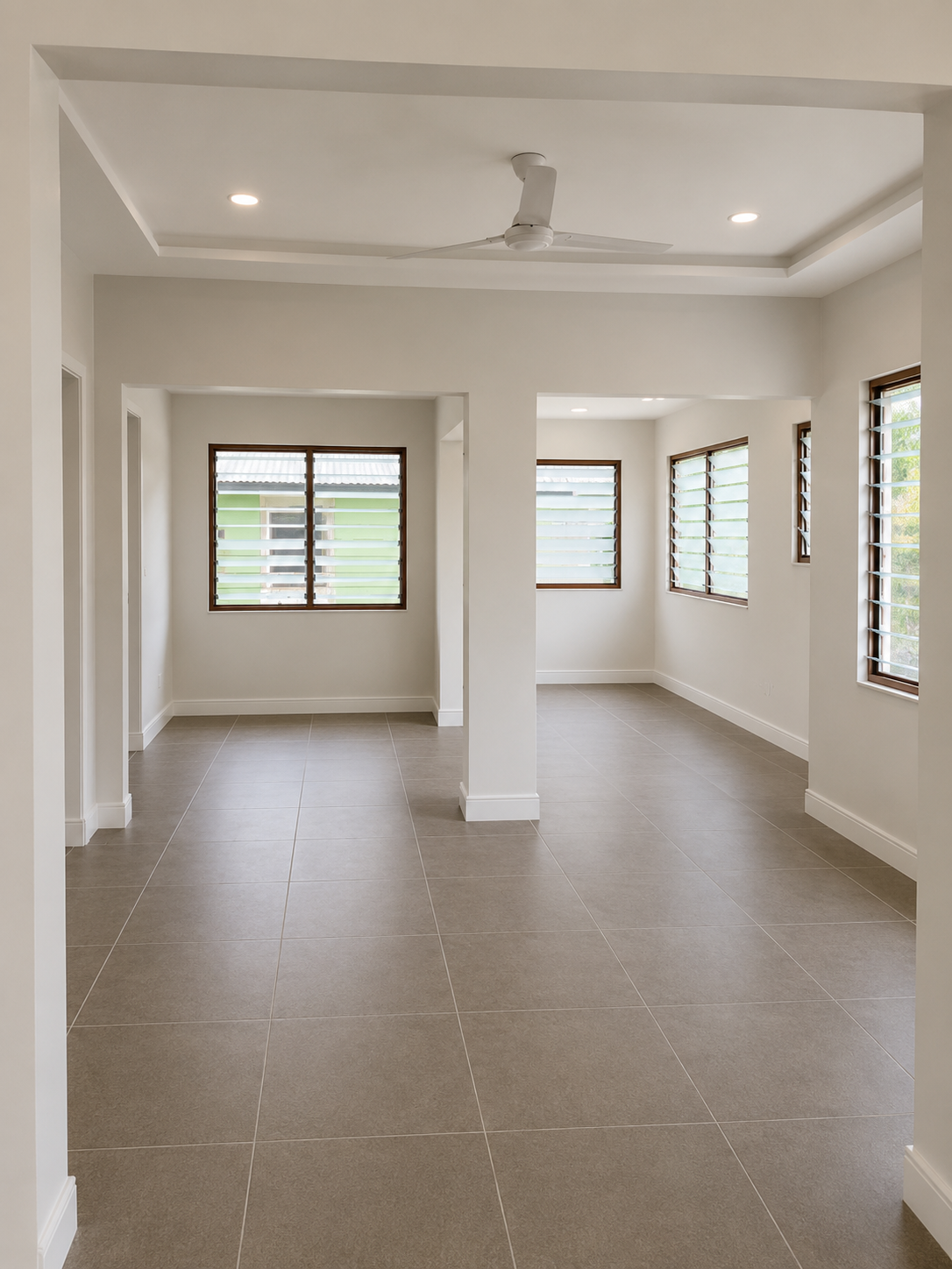 Empty room with beige tiled floor, white walls, multiple windows with wooden louvers, ceiling fan, and recessed lighting.