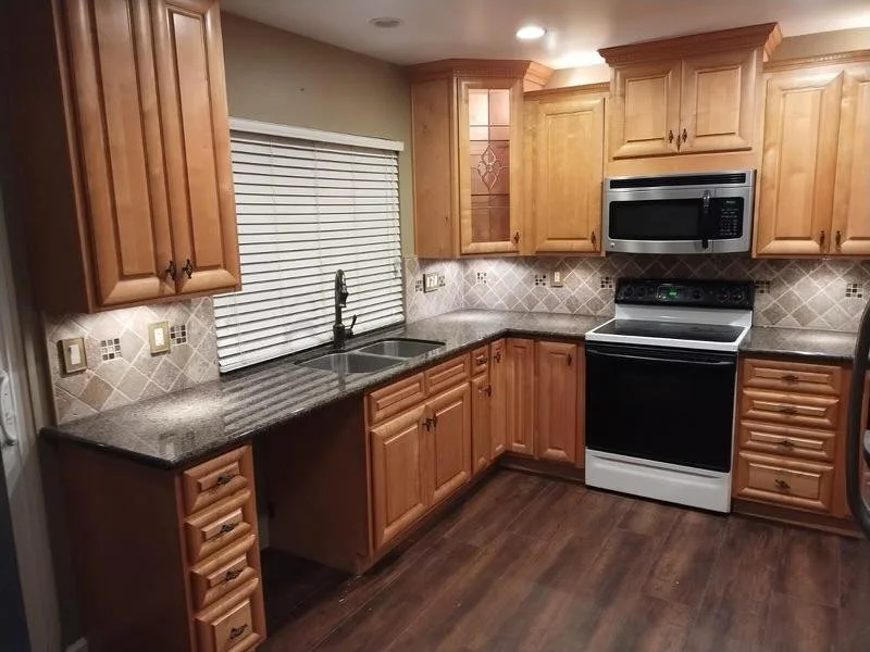 A kitchen with wooden cabinets, a granite countertop, a stainless steel faucet, a sink below a window with closed blinds, a black and white stove with a microwave above, and a tiled backsplash with decorative patterns.