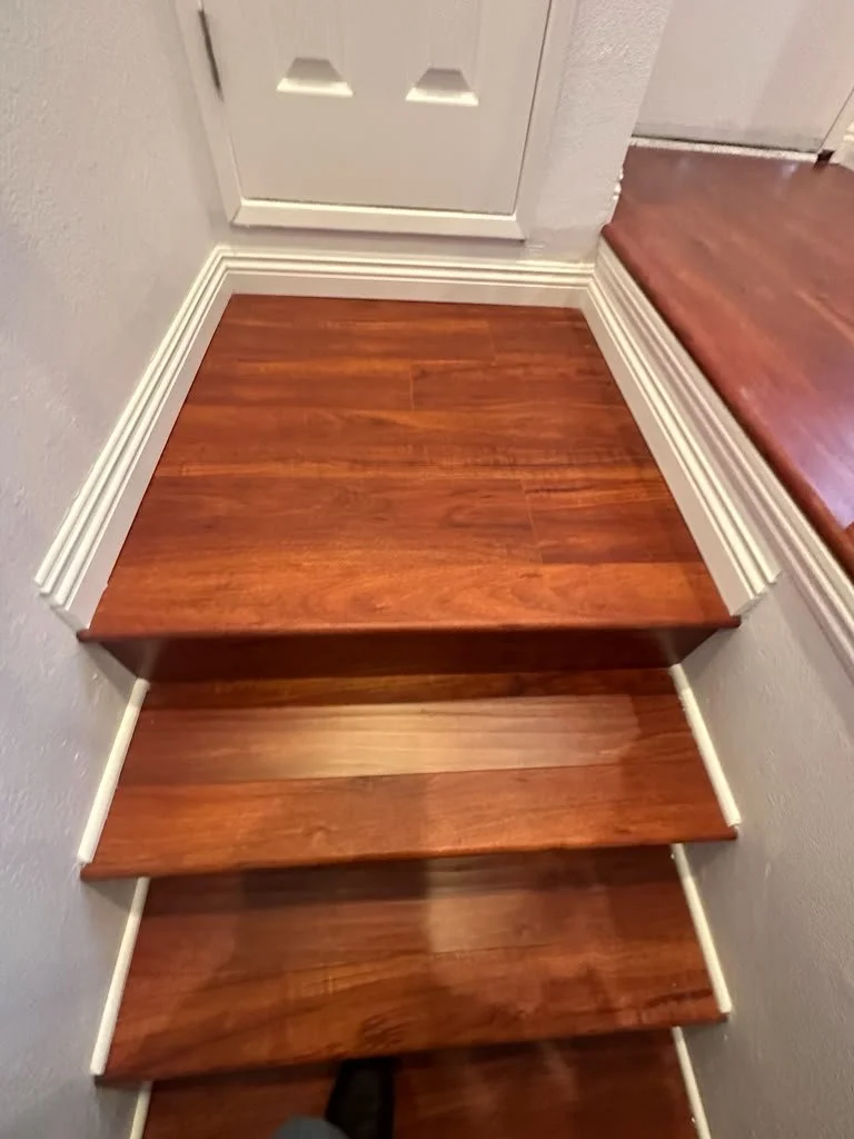 Close-up of wooden stairs with three steps, leading up to a door, with white trim along the edges of the steps and a hardwood floor visible at the top of the staircase.