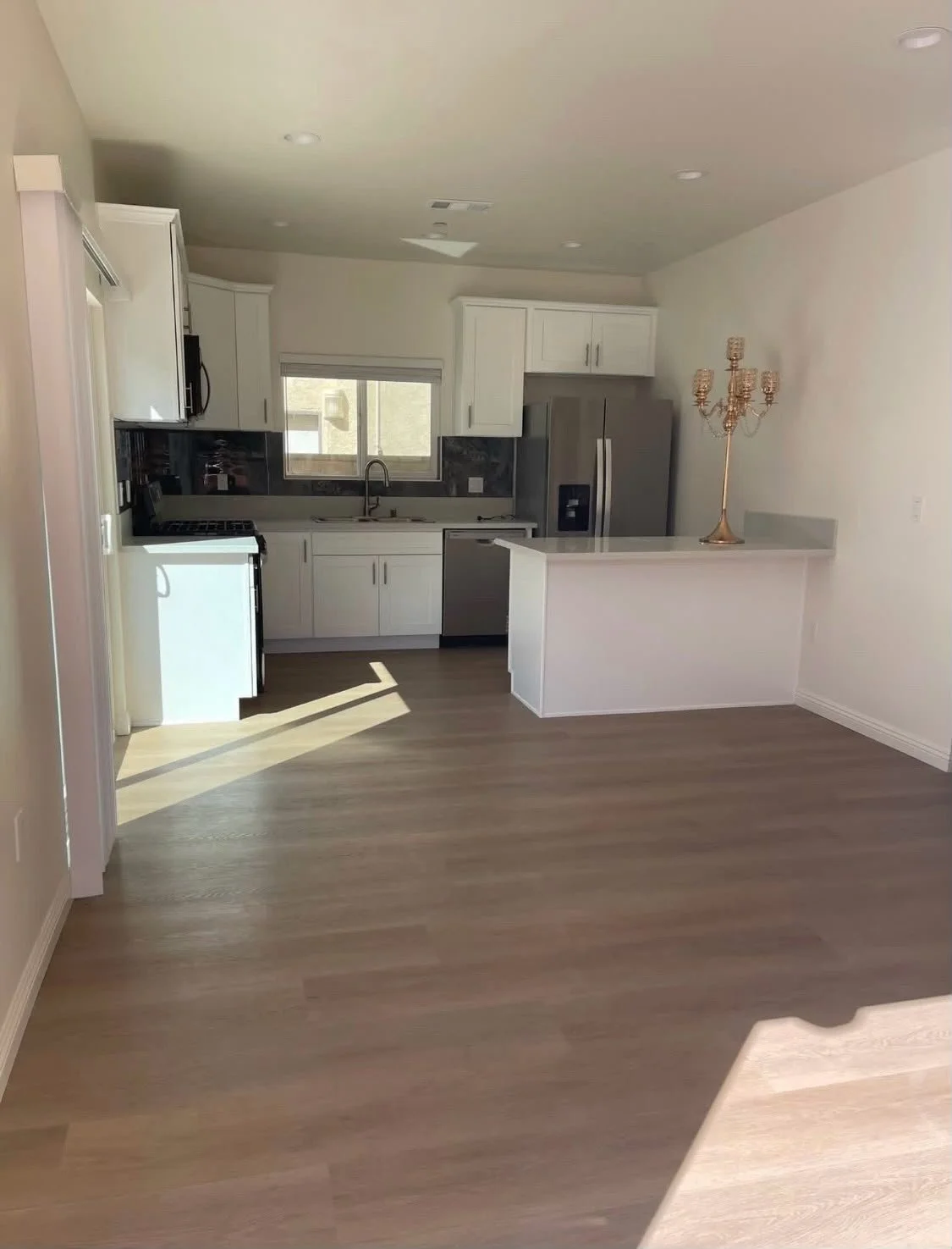 Empty kitchen with white cabinets, stainless steel appliances, and light wood flooring, with sunlight coming through a window.