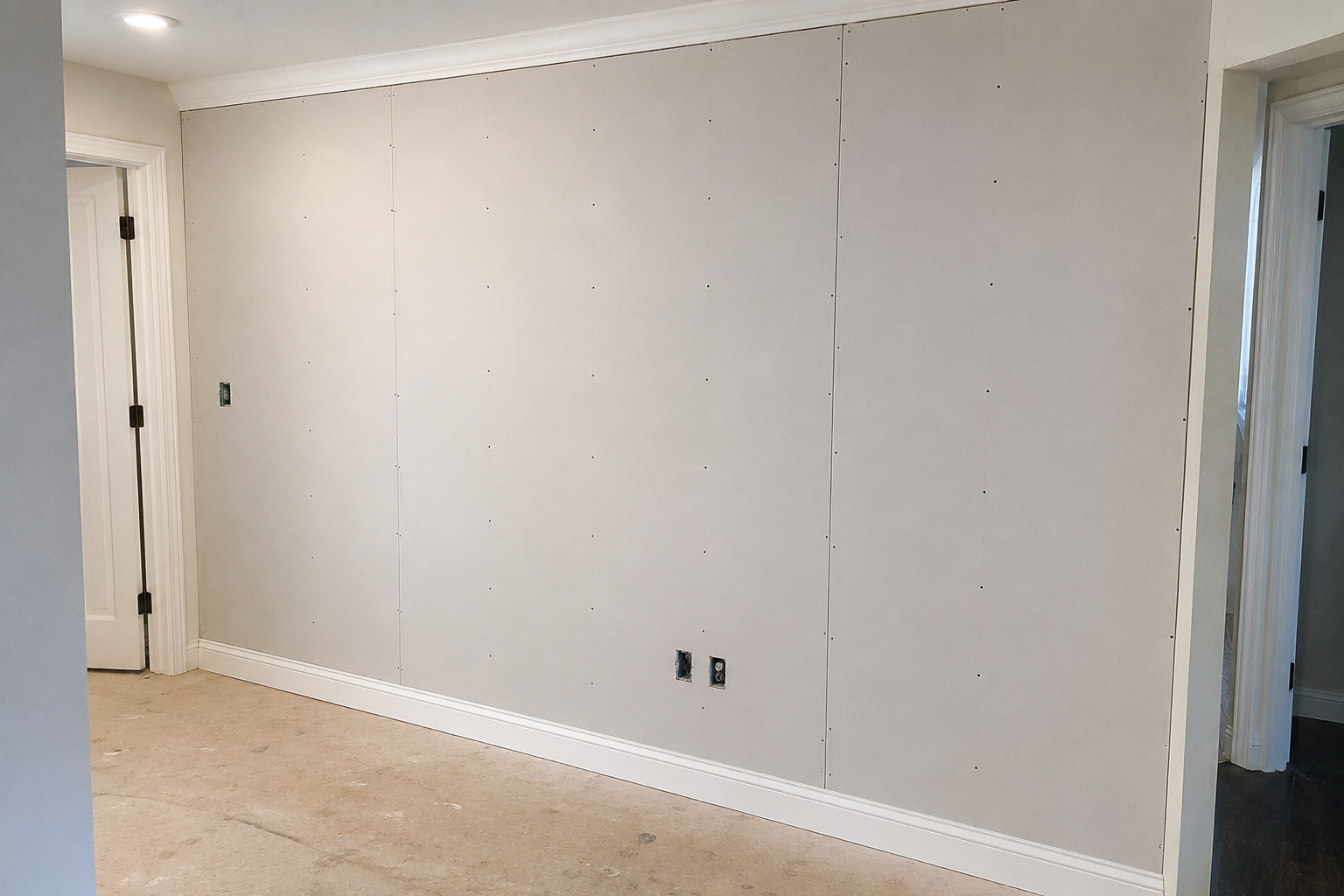 Unfinished interior wall with drywall panels, electrical outlets, and baseboards in a room under construction.