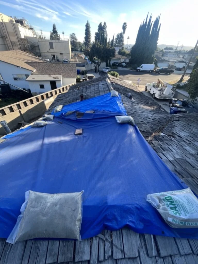 Blue tarp secured on a residential roof for repair or protection, with a bag of washed plastic pellets and a pillow on the tarp.