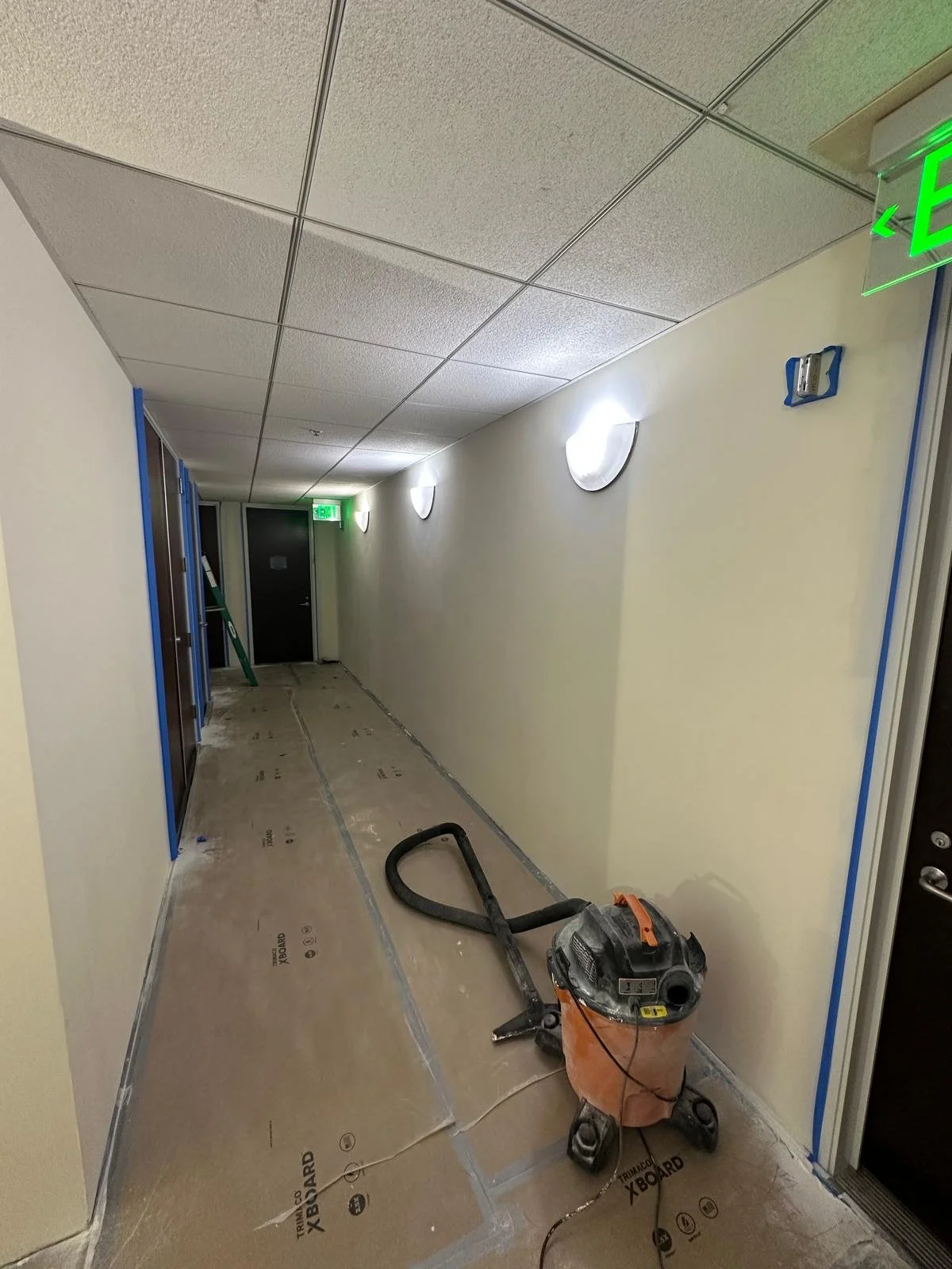 Empty hallway under construction, with protective covering on the floor, a vacuum, several blue-framed openings on the left, a black exit sign on the ceiling, and a closed black door at the end of the corridor.