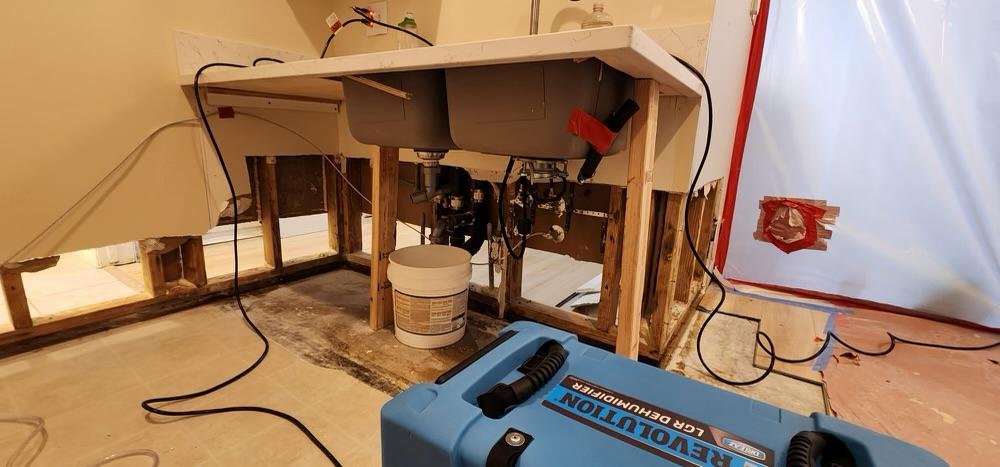 Under the kitchen sink, with exposed pipes and wooden framing, a bucket and plumbing tools are visible. A plumbing machine is situated on the floor nearby, with cords connected to power sources.
