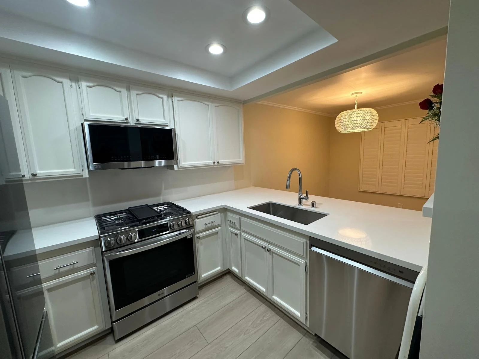 Modern kitchen with white cabinets, stainless steel appliances, a built-in microwave, gas stove, and a white countertop with an undermount sink. Recessed lighting and a patterned hanging light fixture are visible.