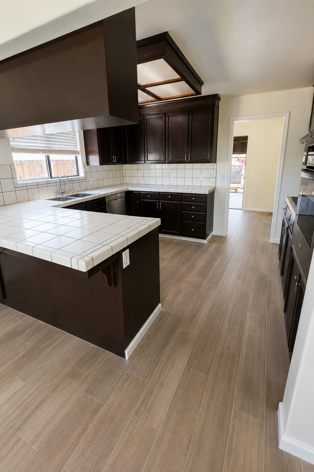 Kitchen with white tiled countertops, dark wooden cabinets, and a white tiled backsplash. Has a window above the sink, a dishwasher, and wall ovens. Light wood flooring and a doorway leading to an outdoor area.