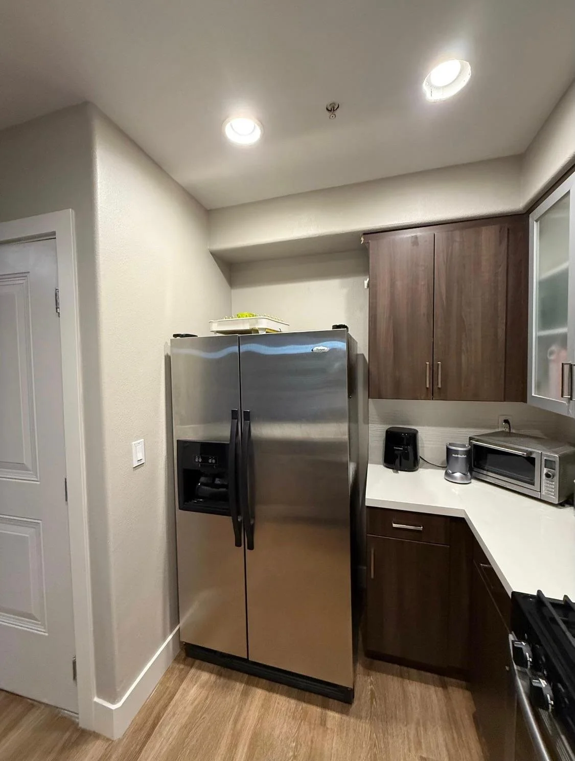 Modern kitchen with stainless steel refrigerator, wooden cabinets, microwave, toaster, coffee maker, and countertop appliances.