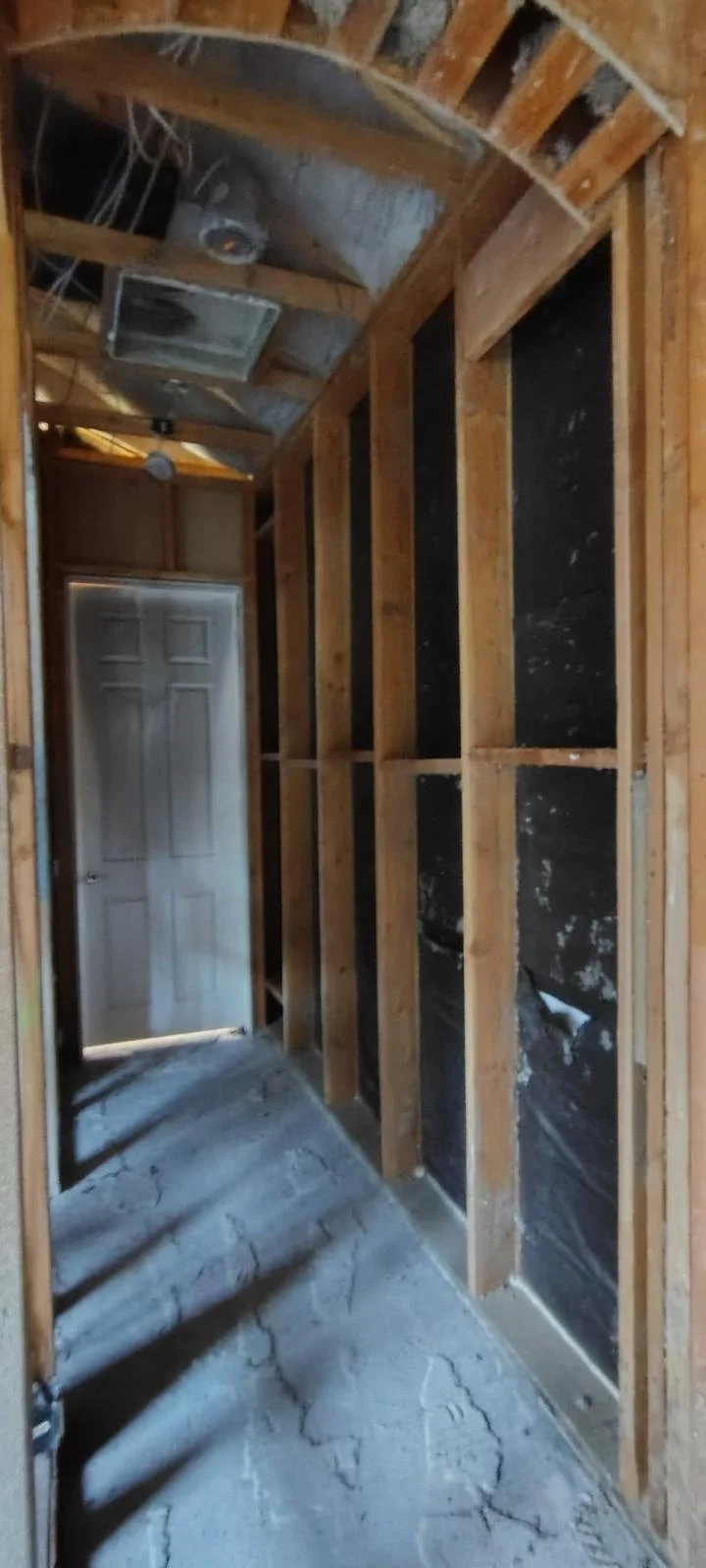 Construction site showing an unfinished room with exposed wooden framing, black insulation on the walls, and a concrete floor with cracks. A door is visible at the end of the room.