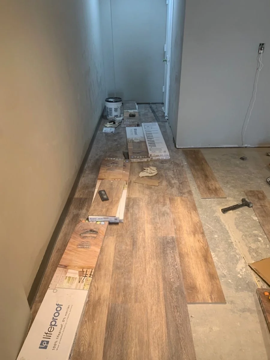 Room under construction with wooden flooring being installed, tools, and flooring boxes on the floor, door at the end.