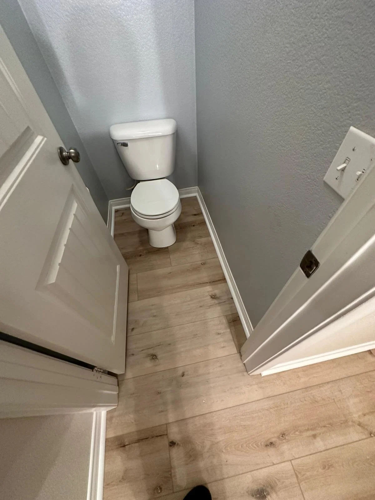 A small bathroom with gray textured walls, a white toilet, wood laminate flooring, a white door, and a light switch on the wall.