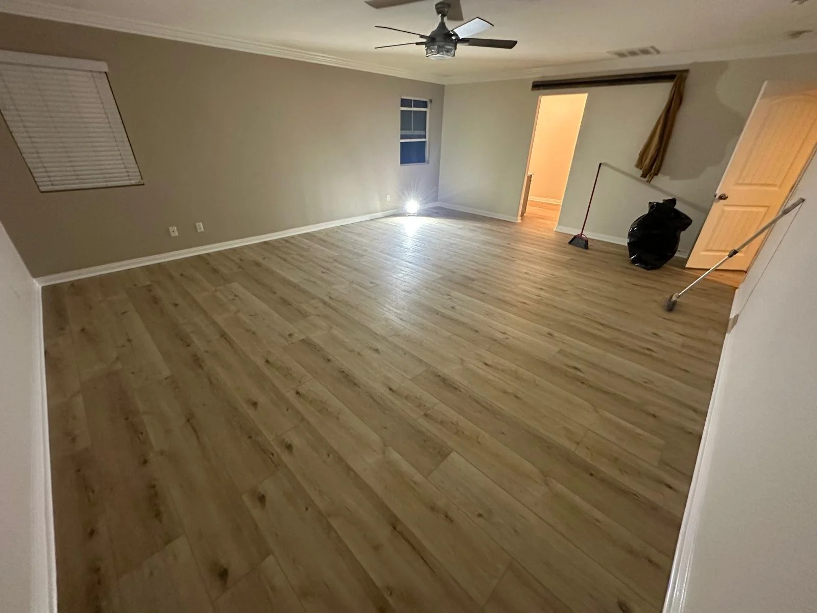 Empty living room with wooden floors, ceiling fan, closed blinds, and a window. Cleaning tools and a trash bag are near an open doorway.