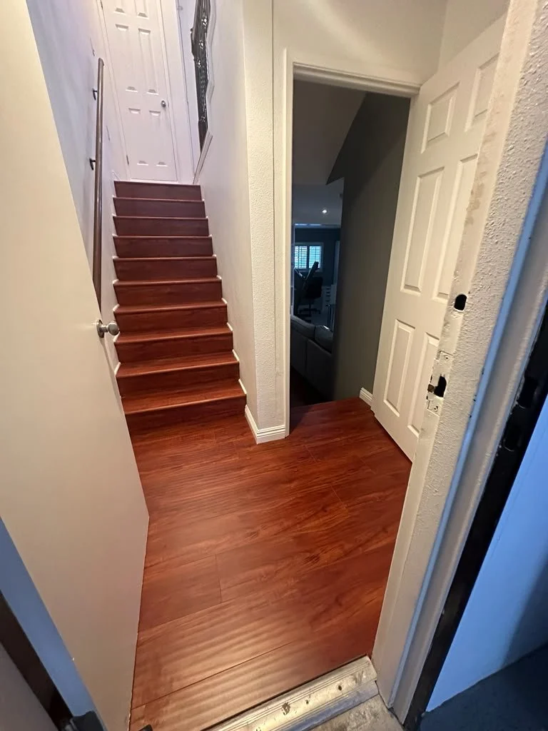 View of a home interior entrance with a staircase leading upstairs, hardwood flooring, and a doorway to a room with a visible couch and windows with blinds.