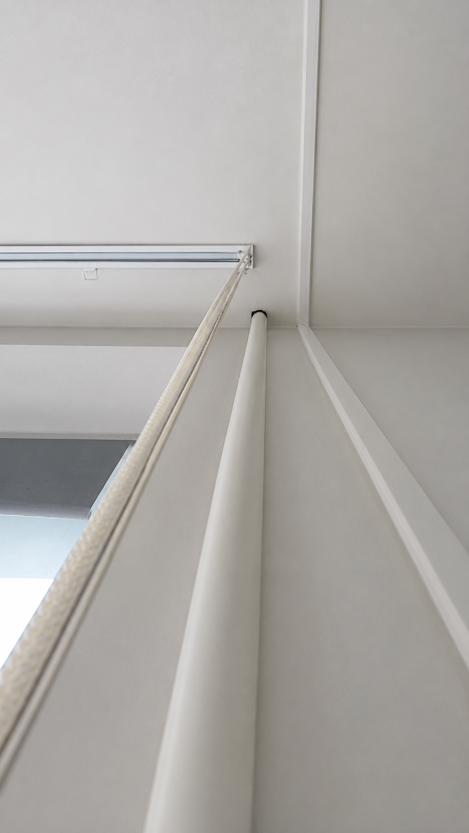 Close-up view of a corner interior ceiling with white walls, a ceiling panel, and a white vertical support or pipe, showing part of a window frame and a blind mechanism.