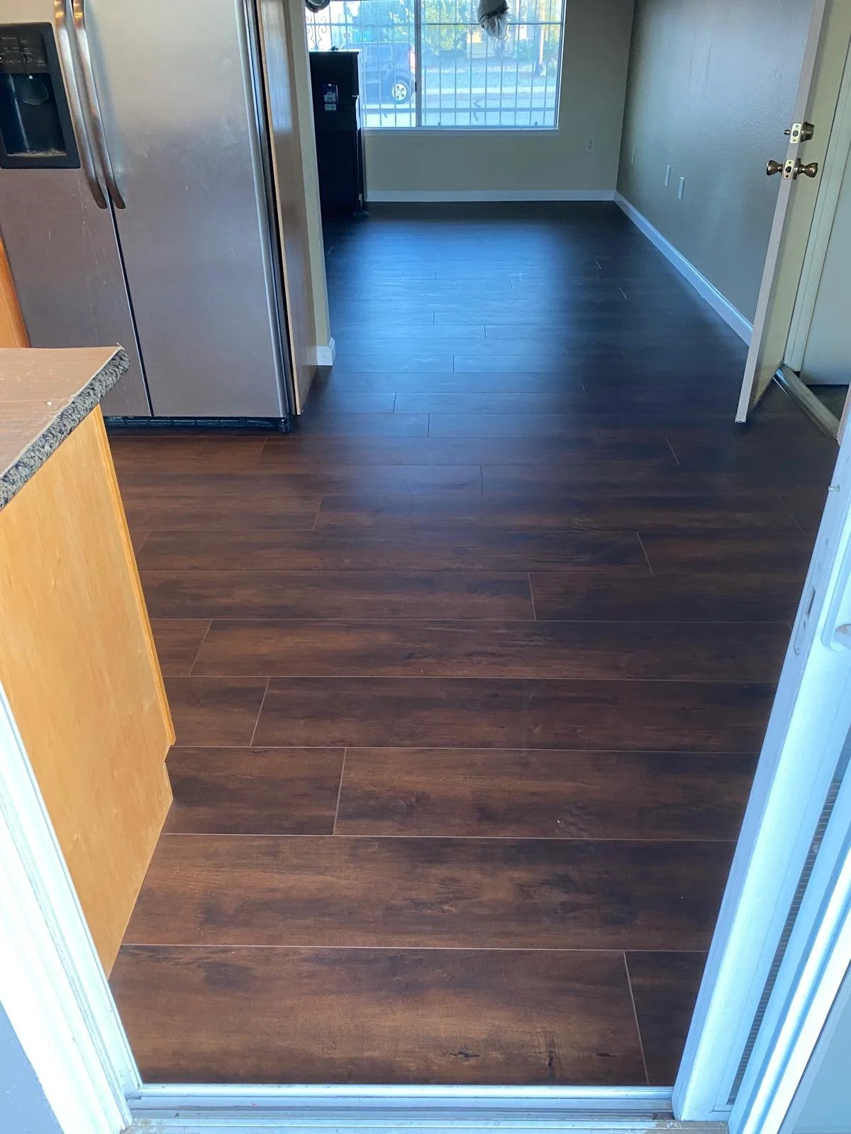 View of a room with dark wood flooring, a window with bars, and a door leading outside. Part of a kitchen counter with a granite surface is visible in the foreground.