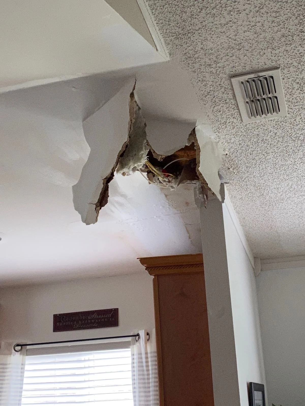 Ceiling with a large hole exposing electrical wiring and drywall debris, near a wall vent and corner of a room, with a window and wooden cabinet below.