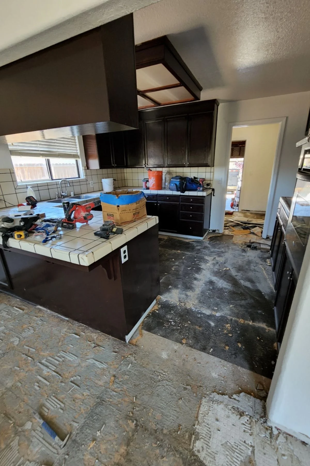Kitchen under renovation with damaged flooring, dark cabinets, tile countertops, various tools, and construction materials scattered around.