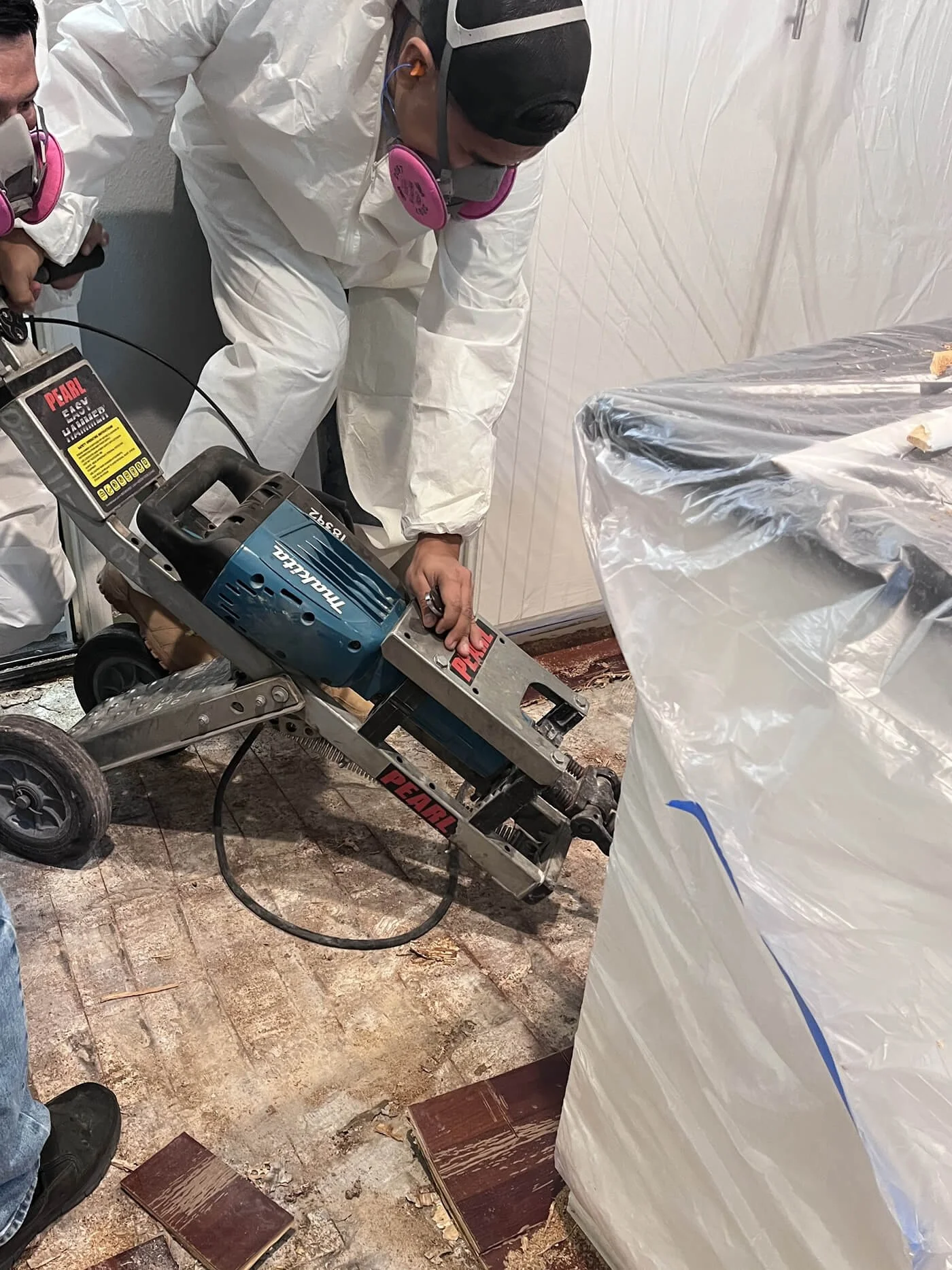 People working in a construction or renovation site, wearing protective masks, white coveralls, and using a power tool to cut or remove flooring material.