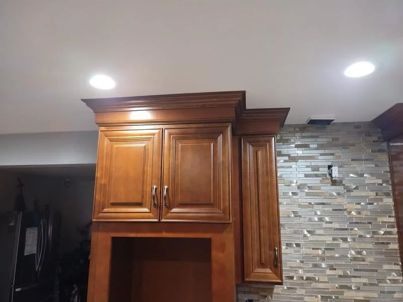 Kitchen ceiling with recessed lighting, wooden cabinets, and a tiled wall with beige and gray tiles.