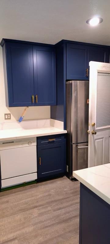 Kitchen with blue cabinets, white countertops, a stainless steel refrigerator, a dishwasher, and a door with blinds.