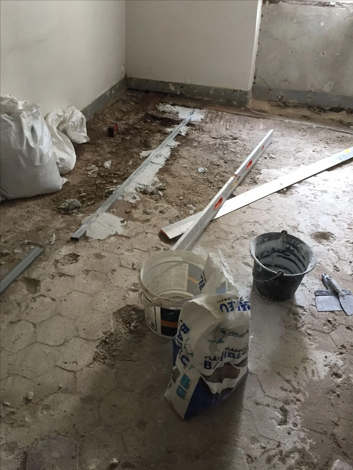 Construction site with dirt floor, tools, and materials, including level, bucket of black paint, and bags of construction supplies, in a room under renovation.