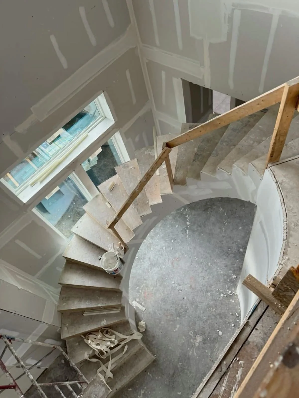 Construction site of a staircase with unfinished drywall and wooden railing, with construction materials and tools around.