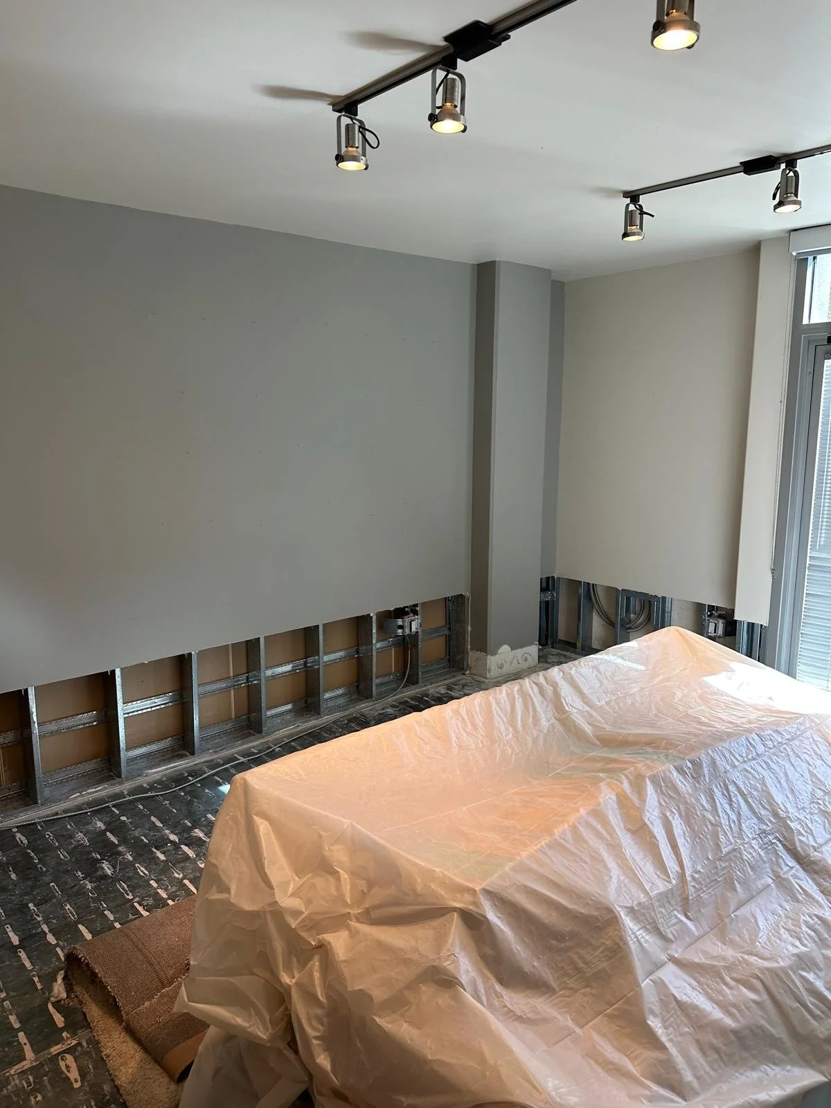 Living room undergoing renovation with exposed wall framing, partially painted walls, a dropped ceiling with track lighting, and a covered piece of furniture