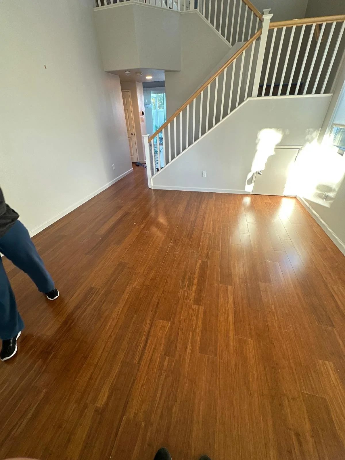 Photo of a living room with hardwood floors, white walls, a staircase with wooden handrails, and a door leading outside. Sunlight is shining through a window, casting shadows on the wall.