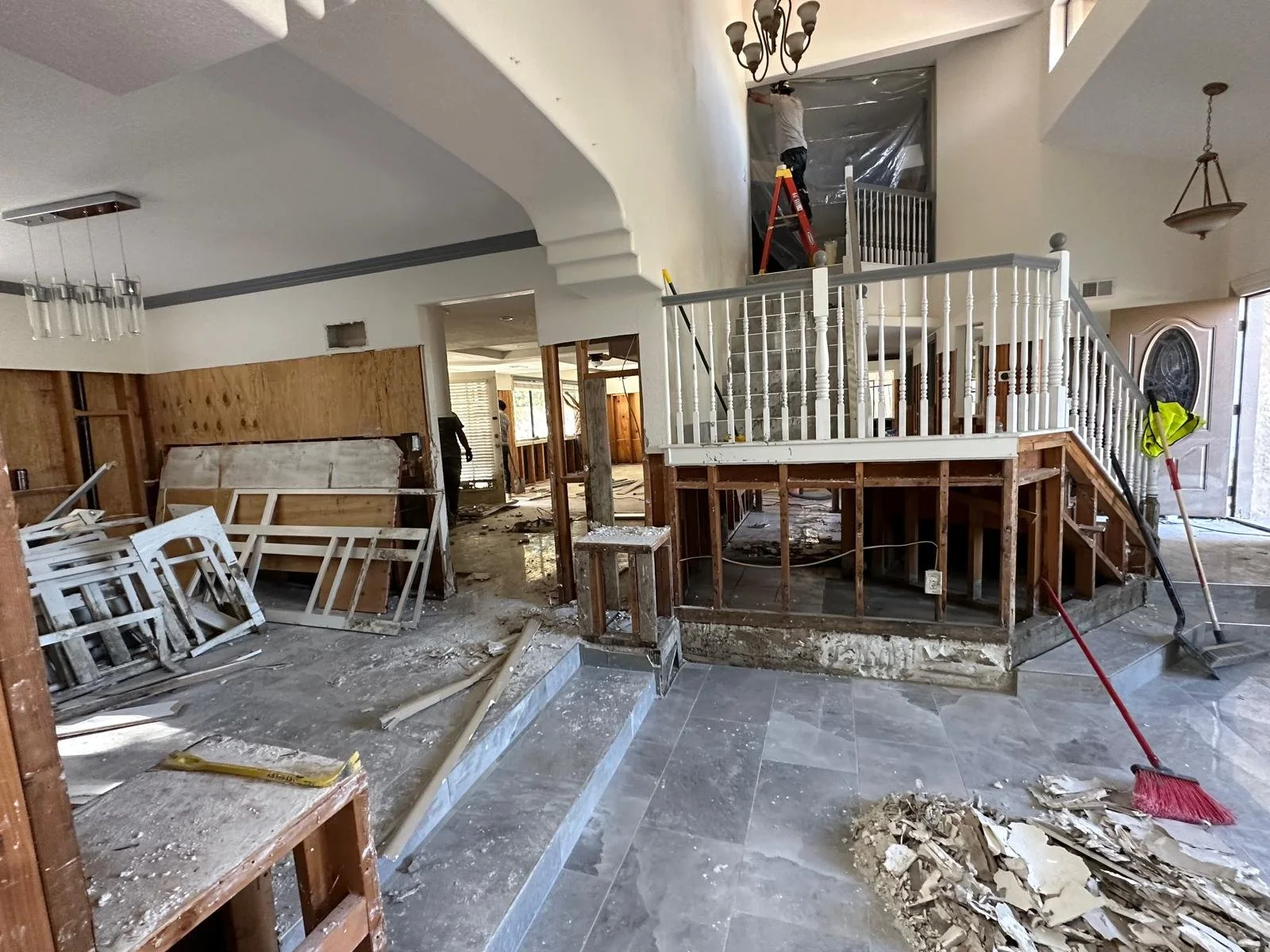 Interior of a house under renovation with exposed wooden framework, scattered debris, and construction tools including a broom and paint roller. A worker is seen on a ladder working on a second-floor opening.