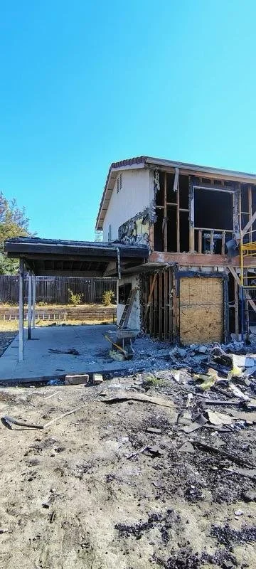A house under construction with exposed framing and partially built walls, construction debris on the ground, and a bright blue sky.