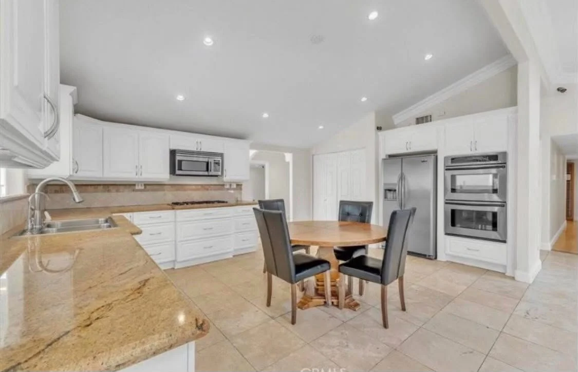 A spacious kitchen with white cabinets, stainless steel appliances, and a round wooden dining table with four black cushioned chairs, beige tiled floor, and recessed ceiling lights.
