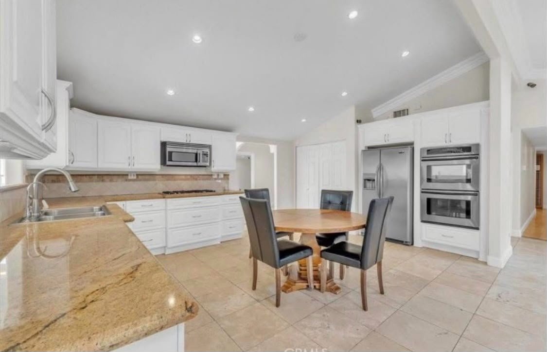 A spacious kitchen with white cabinets, stainless steel appliances, and a round wooden dining table with four black cushioned chairs, beige tiled floor, and recessed ceiling lights.