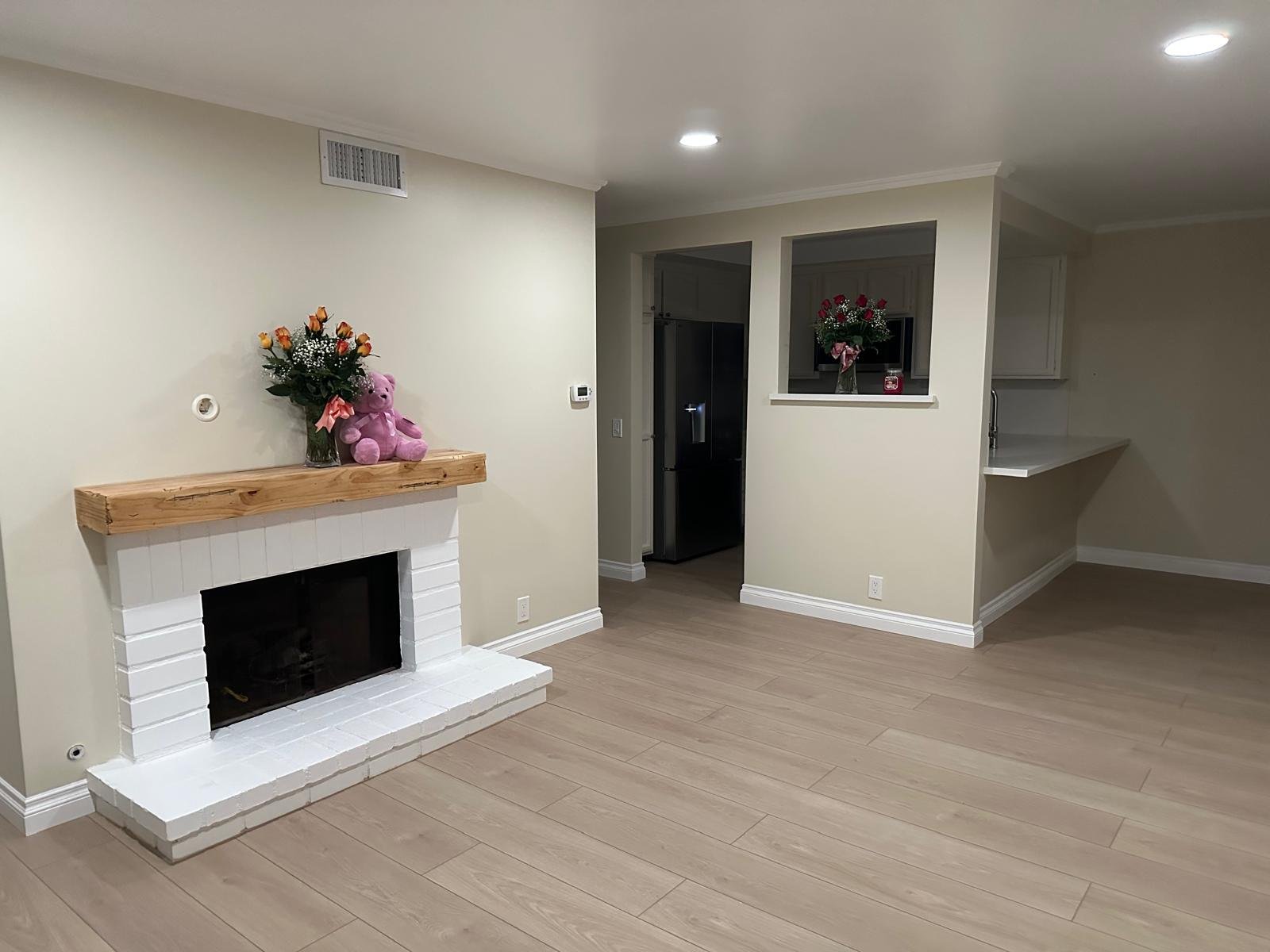 Empty living room with a white painted fireplace, decorated with a pink teddy bear and a flower arrangement, beige walls, and light-colored wooden flooring.