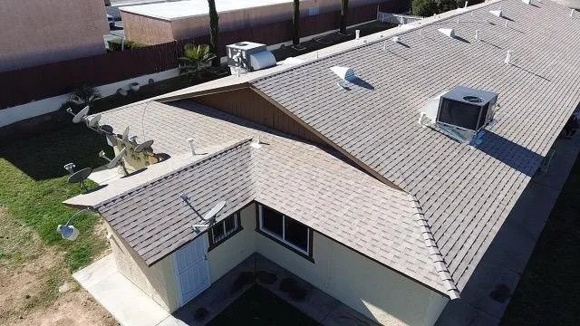 Aerial view of a residential house with a gray roof and white exterior walls, showing appliances and vents on the roof and a small yard with a grassy area and concrete walkway.