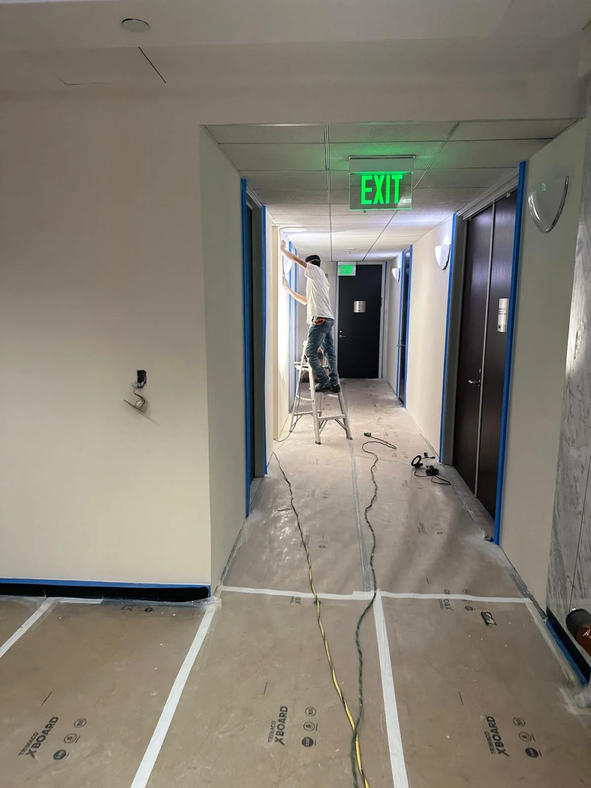 A worker standing on a ladder installing or repairing an electrical or data cable in a hallway with multiple doorways, with a green illuminated 'EXIT' sign overhead.