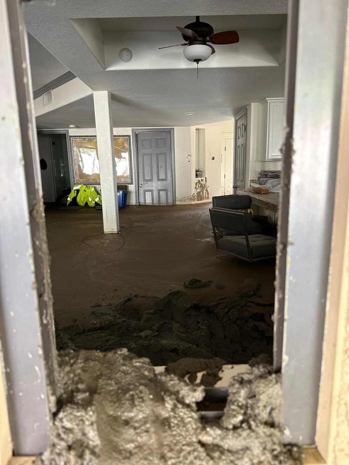 Photo of a house interior seen through a partially broken wall, showing a room with dirt and mud on the floor, a ceiling fan, and some furniture like chairs and a counter.
