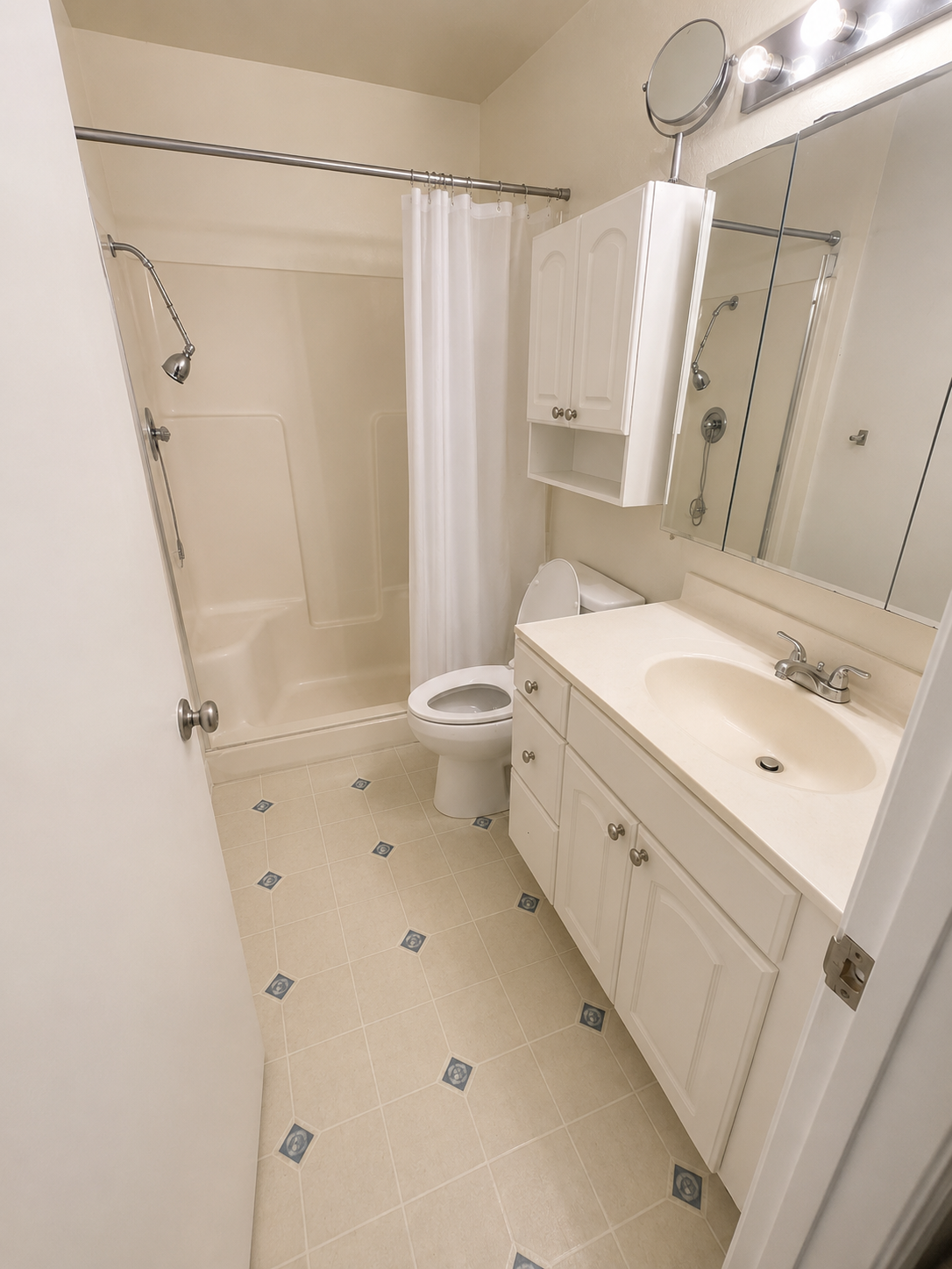 Small bathroom with beige tiles, a white shower stall with a white curtain, a toilet, a white vanity with a sink, a large mirror, and a wall-mounted cabinet.