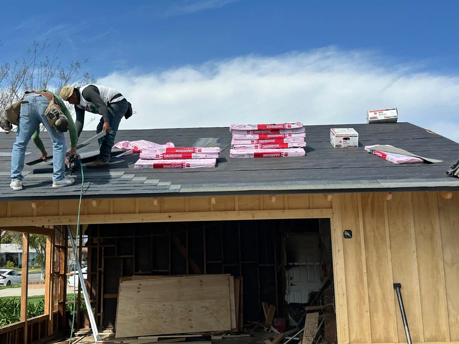 Two construction workers installing roofing shingles on a house under a blue sky, with stacks of roofing materials nearby.