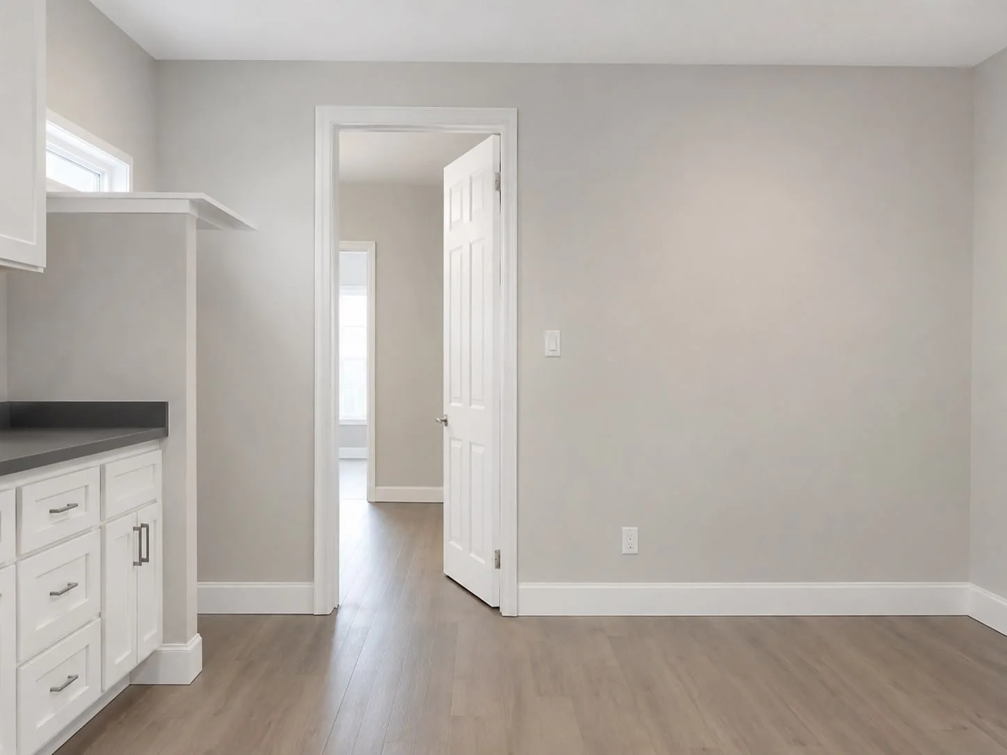 Empty room with gray walls, white trim, and hardwood floors. There is a doorway leading to another room with a window, and part of a kitchen countertop with white cabinets is visible on the left.