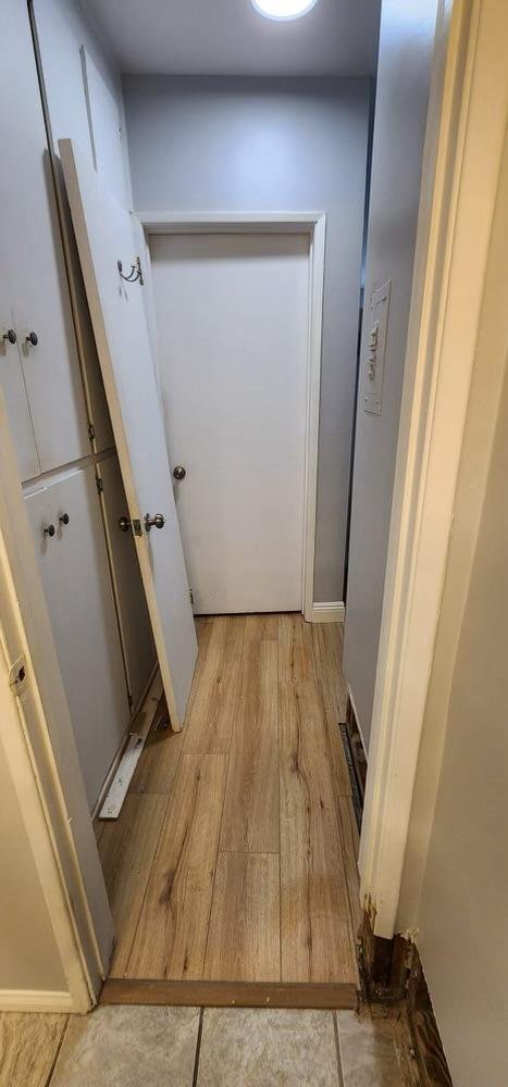 A narrow hallway with a small white door at the end, light gray walls, a wooden floor, and a partial view of cabinets on the left side. One cabinet door is leaning against a cabinet, and there's an air vent on the right floor near the wall.