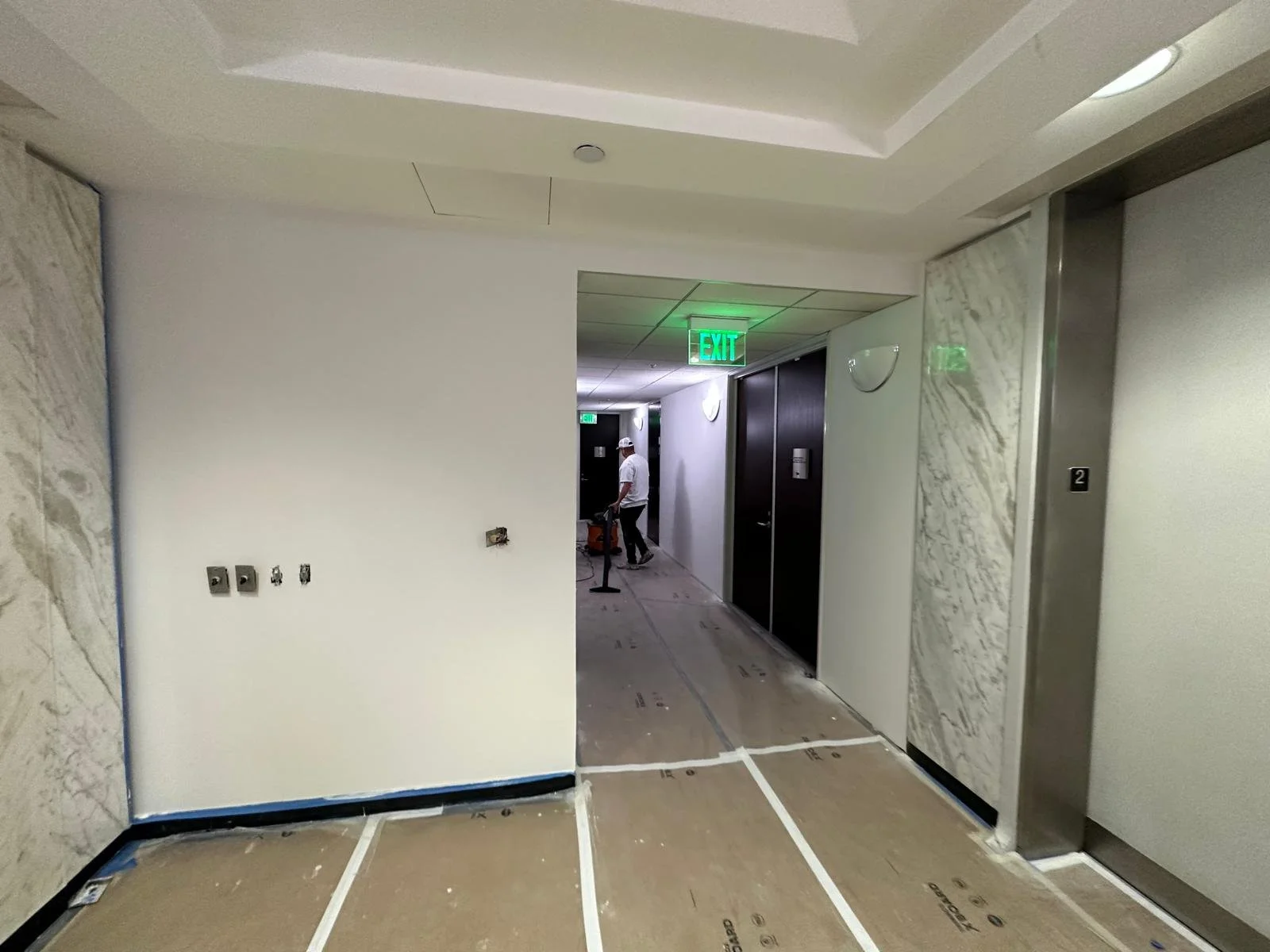 Interior hallway under construction with an elevator on the right, white walls, marble accents, and a worker in the distance near an exit sign.