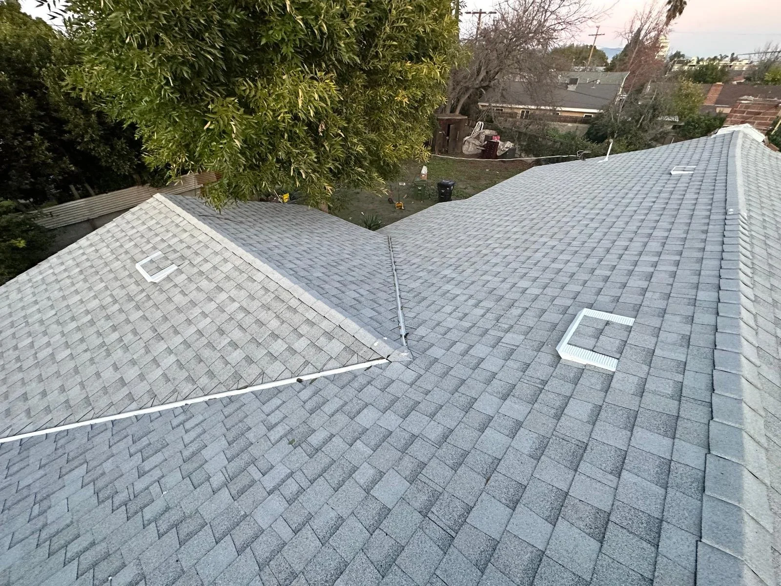 Aerial view of a house roof with grey shingles and rooftop vents, surrounded by trees and neighboring houses.