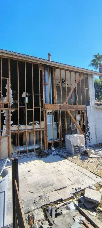 Partially demolished house exterior with exposed wooden framing, damaged wall, and an outdoor air conditioning unit under a clear blue sky.