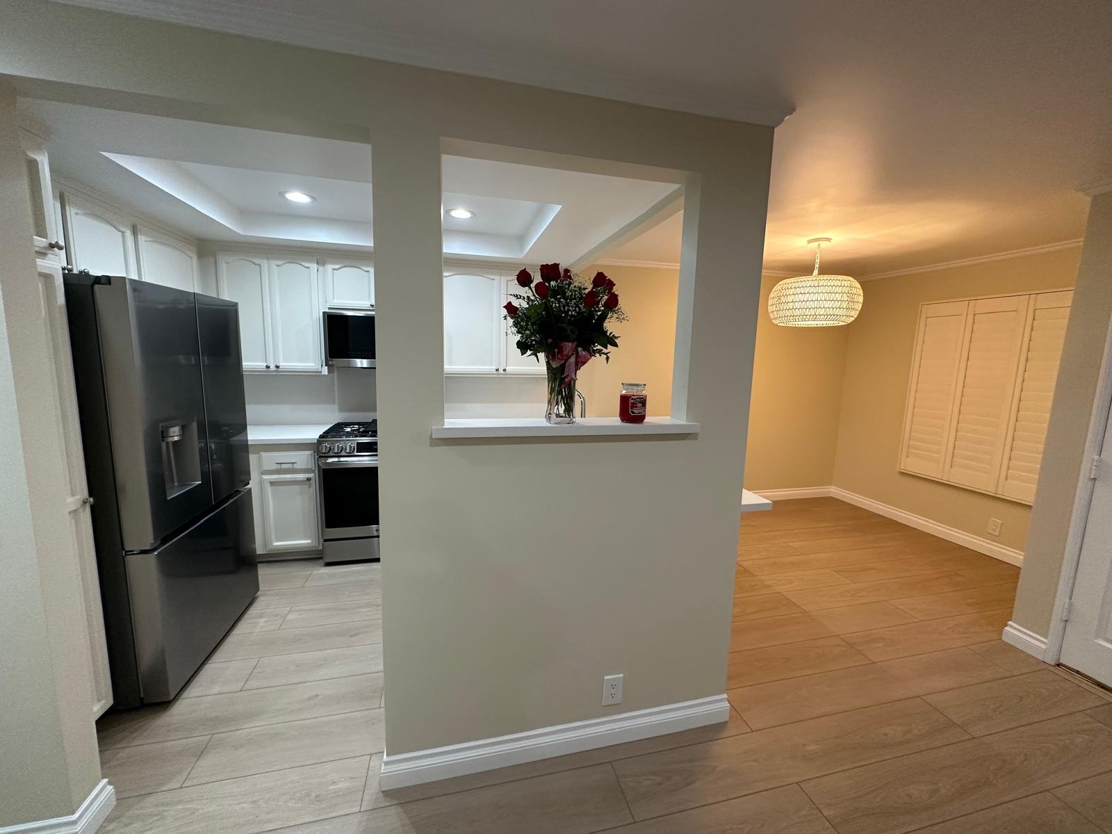 Kitchen with white cabinets, stainless steel refrigerator, black stove, and a vase of red roses on a half-wall. Adjacent room with a beige wall, closed window shutters, and a spherical ceiling light.