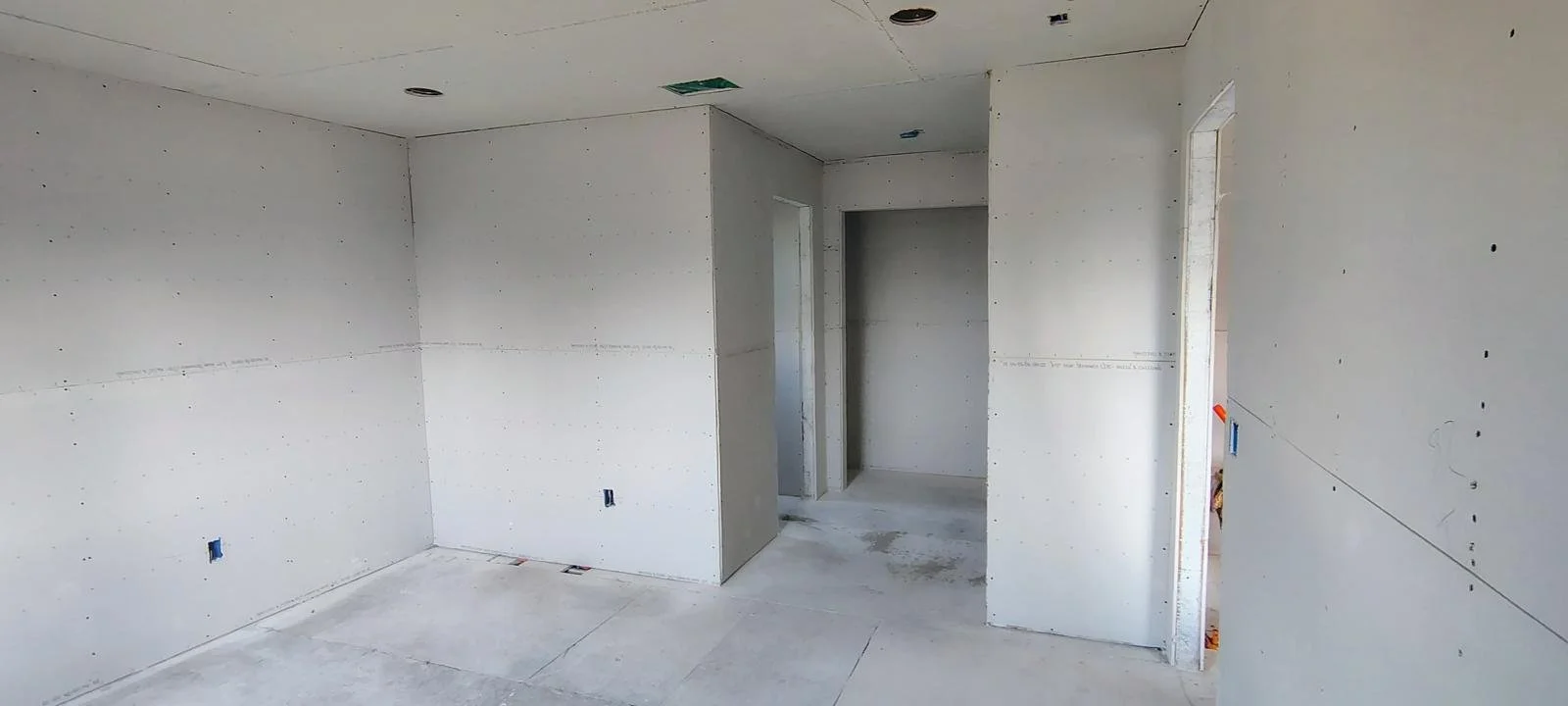 Interior of a room under construction with unfinished drywall walls and floors, with electrical outlets and ceiling fixtures installed.