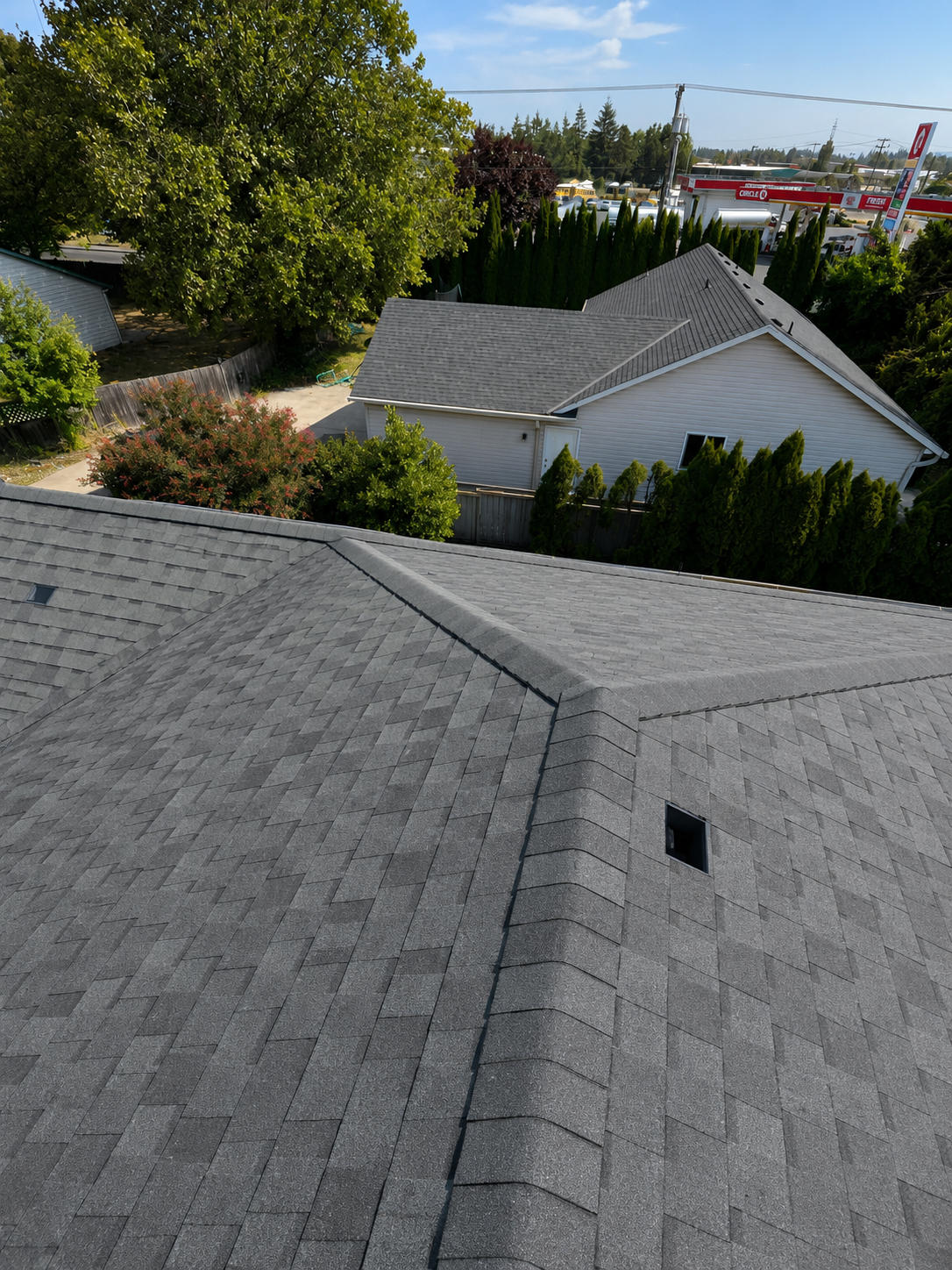 View of rooftops with gray shingles, surrounding trees, and a backyard with bushes and a small shed, with a commercial area in the distance.