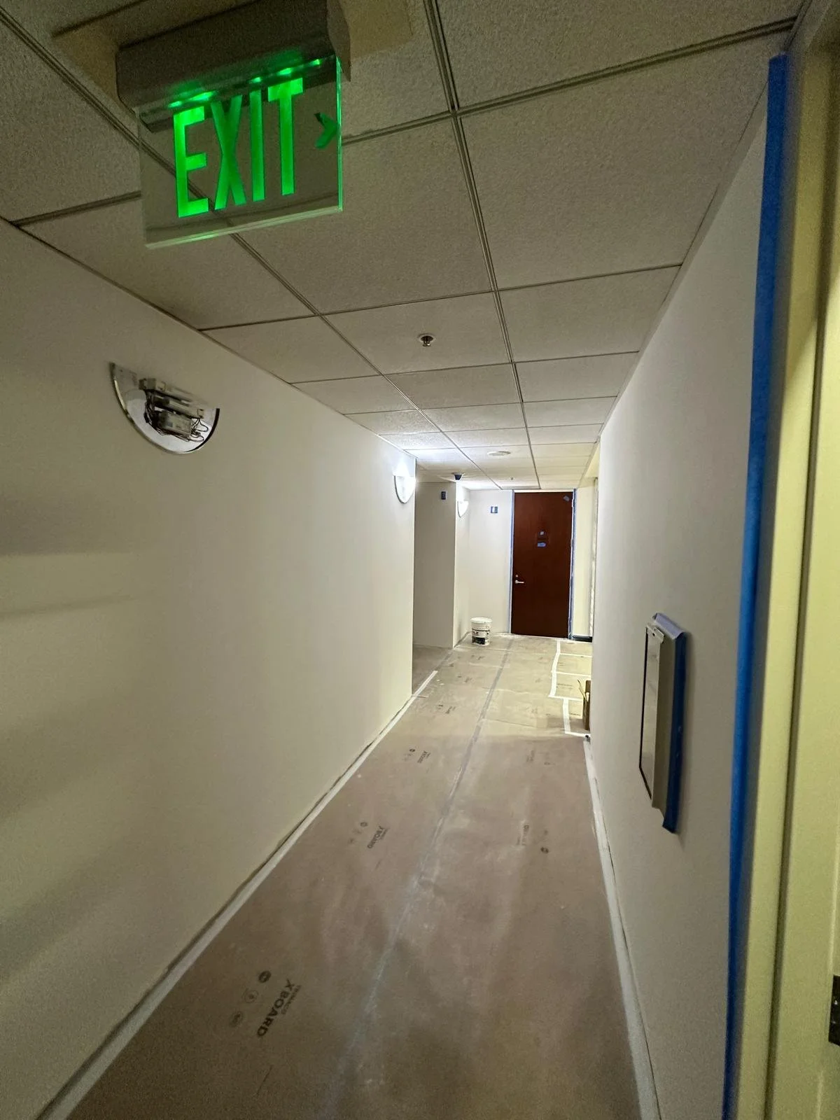 Hallway under construction with a green illuminated exit sign on the ceiling, plain white walls, a closed wooden door at the end, and construction materials on the floor.
