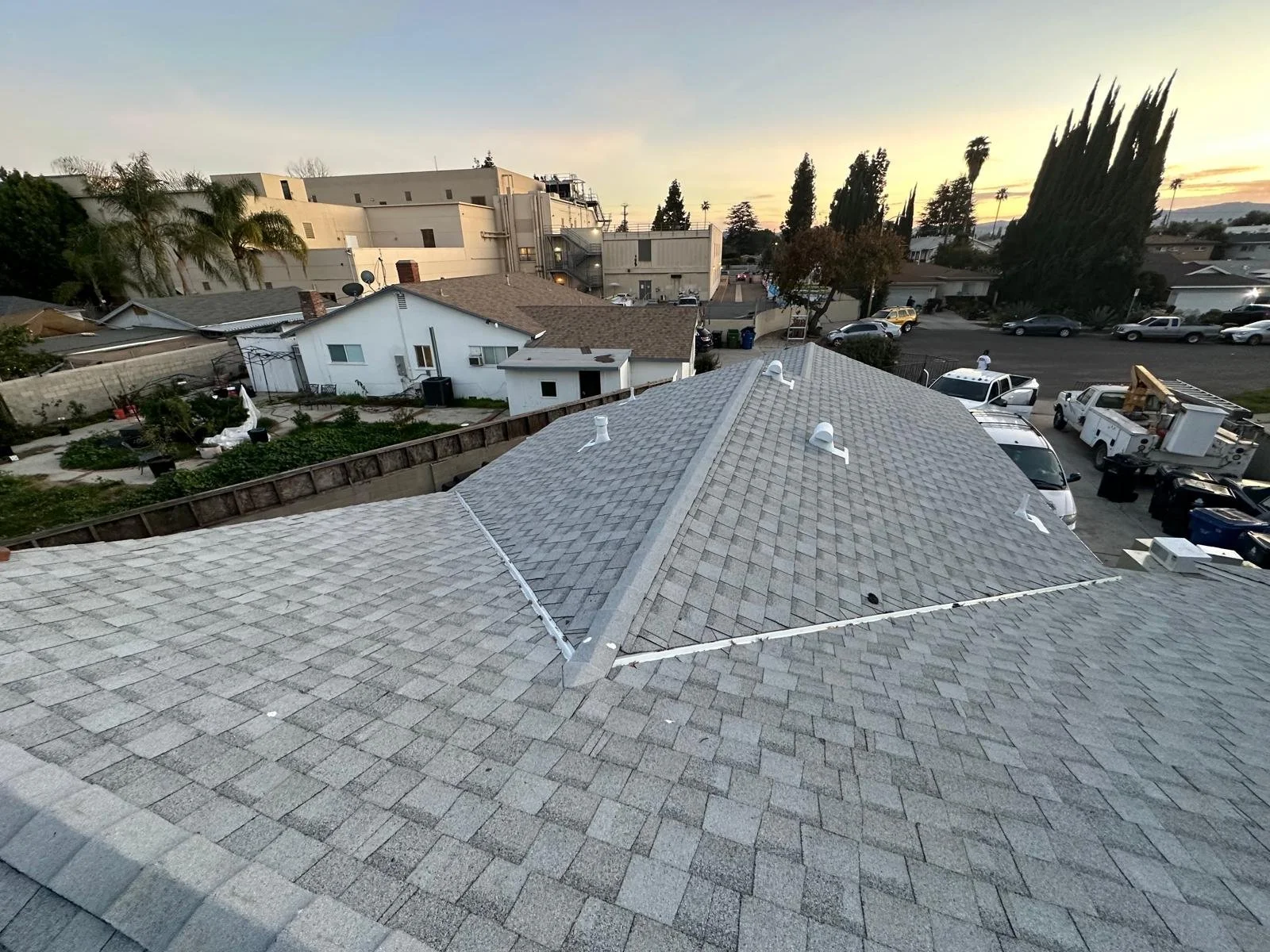 View of a neighborhood and parking lot taken from a rooftop at sunset, showing neighboring rooftops, trees, and buildings.