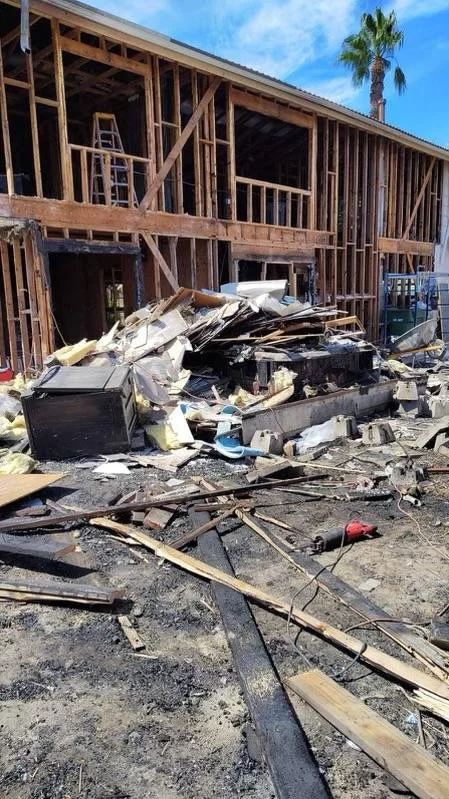 Construction site with damaged building and scattered debris, including broken furniture and burnt materials, under a clear sky with a palm tree in the background.